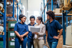 Female manager discussing delivery schedules with staff in warehouse. Female supervisor talking with employees in warehouse.