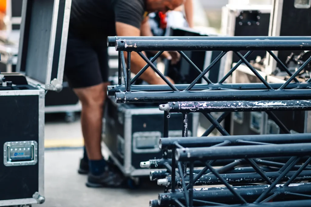 Event production crew member loading stage truss and equipment cases during event setup, with stacked black truss sections in the foreground and road cases in the background.