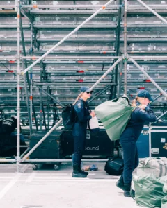 Two crew members in navy uniforms and caps unload green duffel bags in front of a scaffolding grandstand, next to black road cases marked "sound."