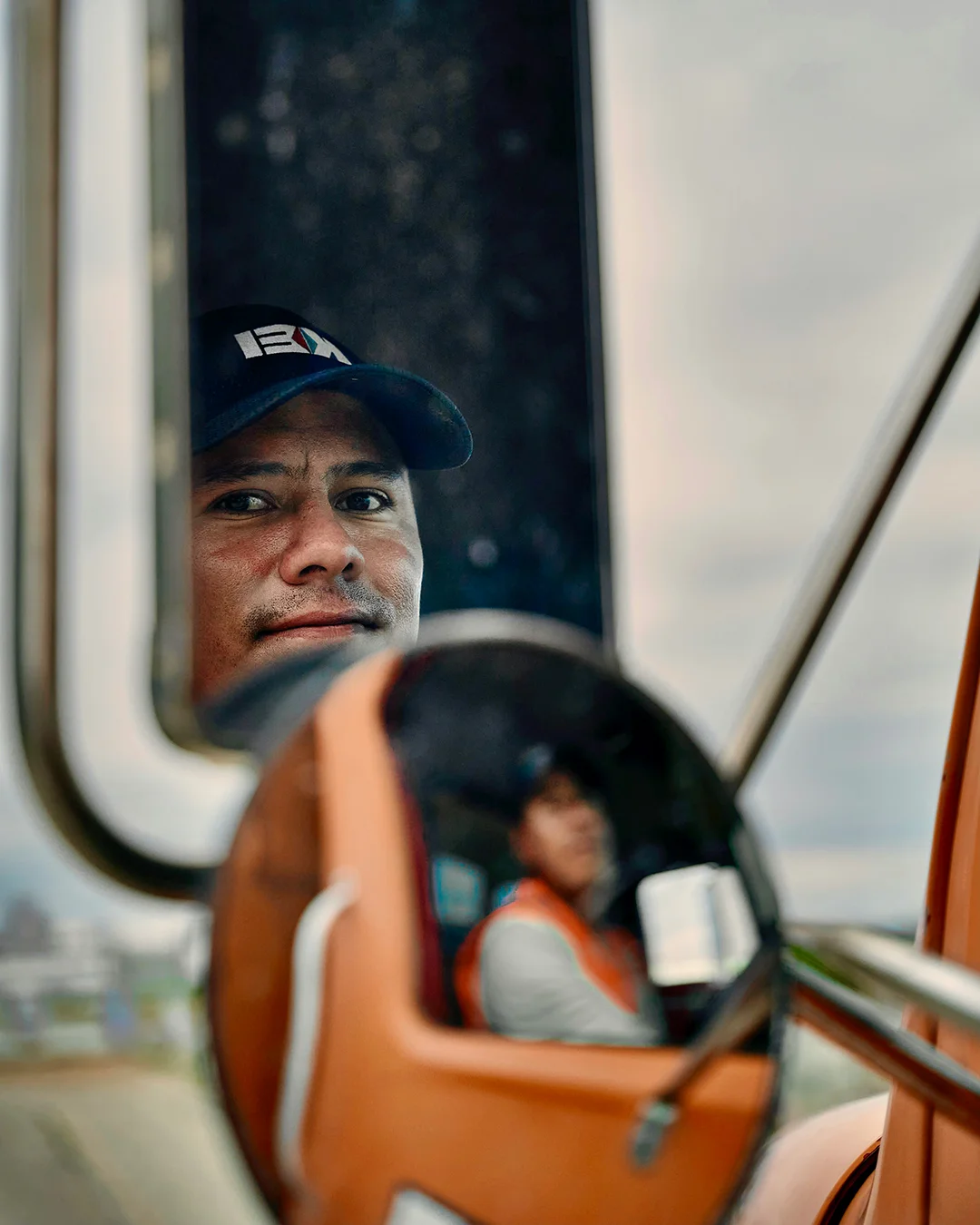 Event equipment operator in a navy cap looks over a vehicle's side mirror, while the mirror shows him seated in the cab wearing an orange safety vest.